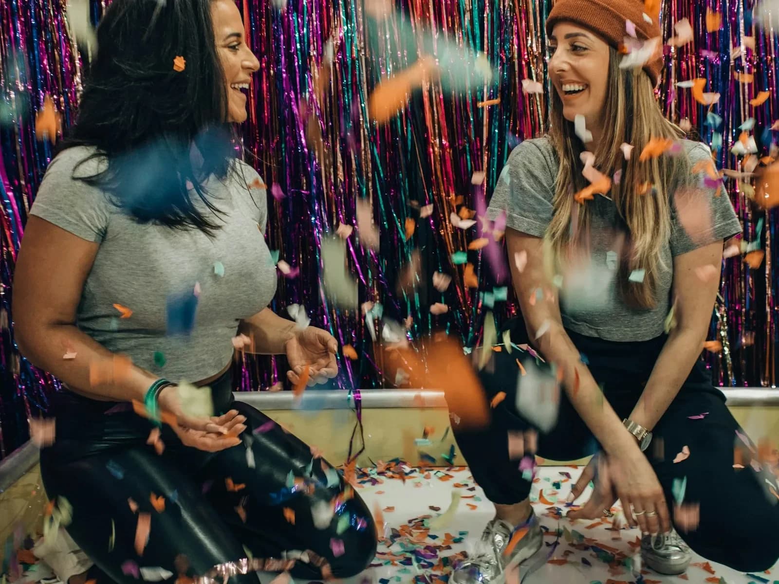 Two women kneeling and laughing together under falling confetti