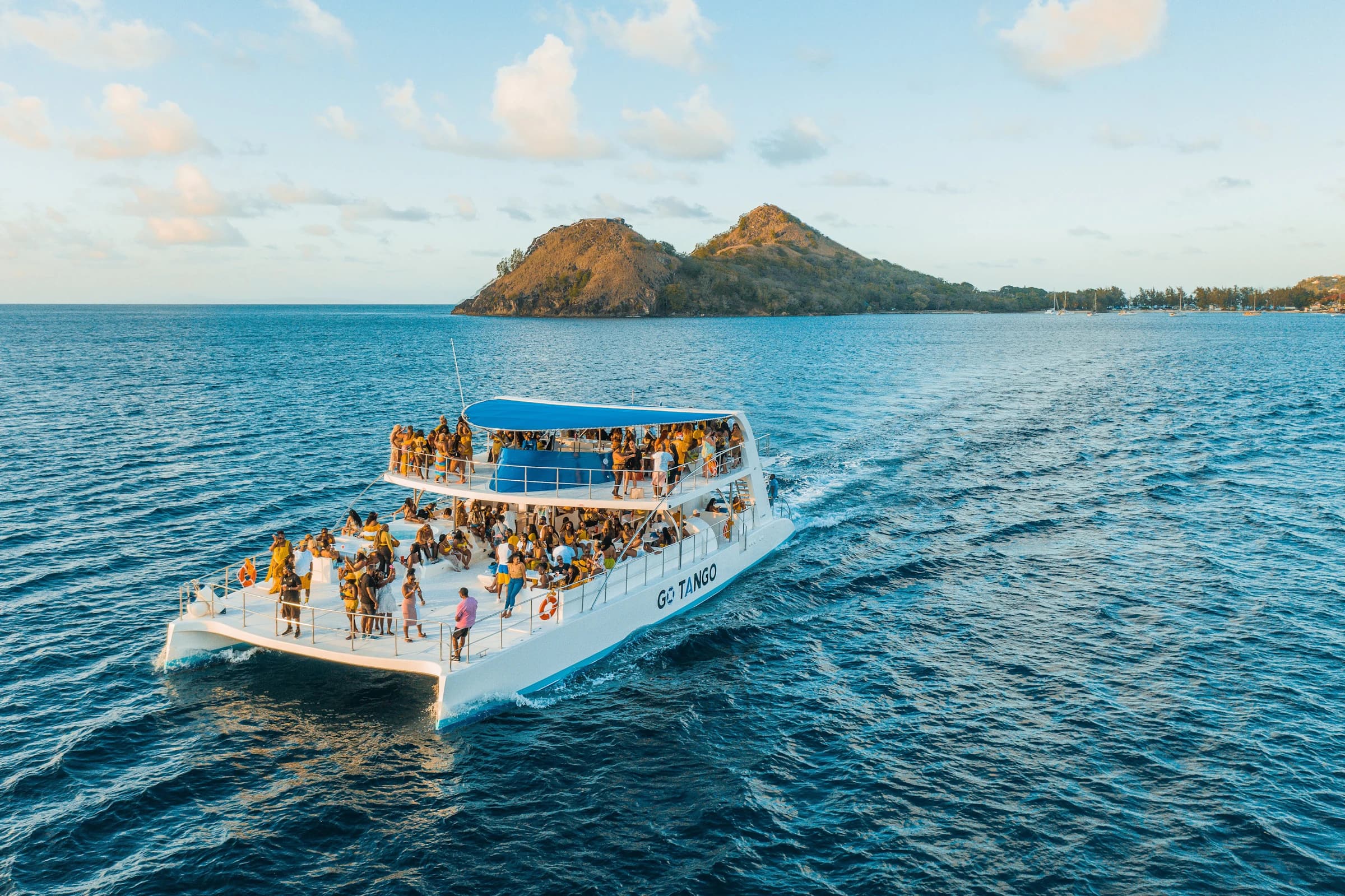 A crowded double-decker party boat cruising past a tropical island at sunset